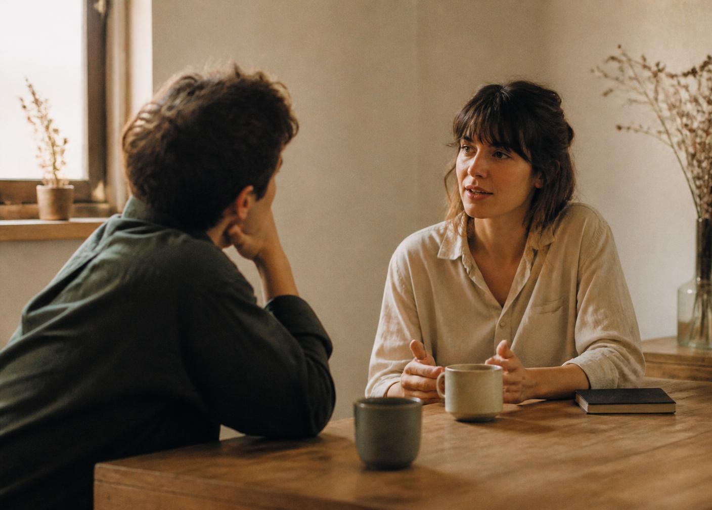 Two people in an unhurried conversation across a wooden table in a quiet room with natural light